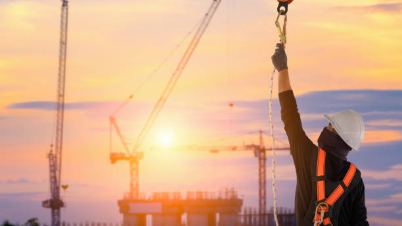 Construction worker wearing personal fall protection harness attaching a snap hook to a pole on the construction site. The sun is setting in the background.