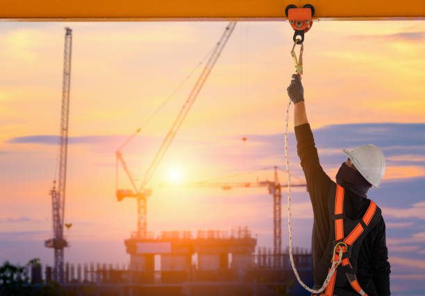 Construction worker wearing personal fall protection harness attaching a snap hook to a pole on the construction site. The sun is setting in the background.
