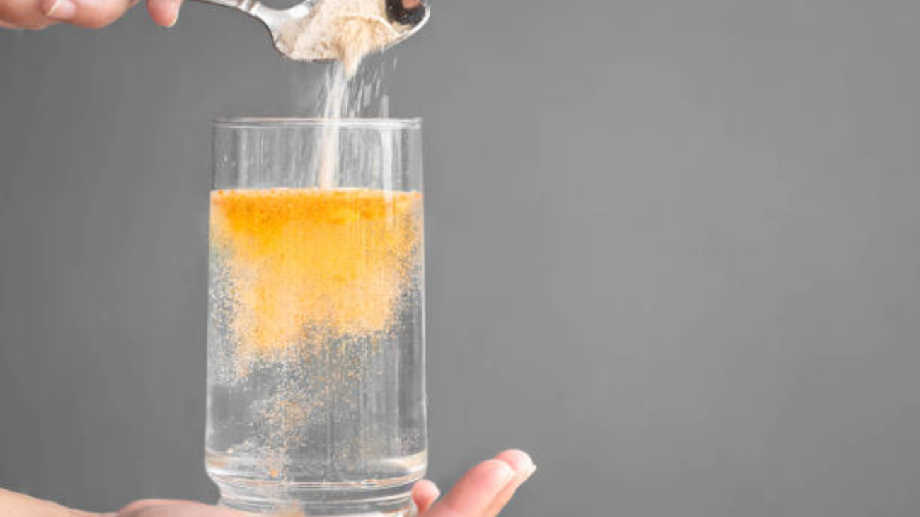 A hand holds a clear glass filled with water, the other hand holds a spoon that is pouring orange electrolyte powder into the water.
