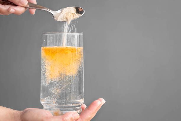 A hand holds a clear glass filled with water, the other hand holds a spoon that is pouring orange electrolyte powder into the water.