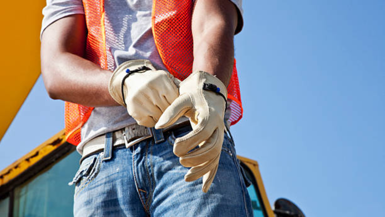 Low angle, cropped view of an African American young man putting on work gloves. He is a construciton worker, wearing an orange safety vest and jeans. Part of the crane is visible behind him, against the clear, blue sky.