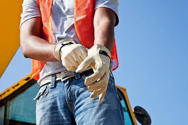 Low angle, cropped view of an African American young man putting on work gloves. He is a construciton worker, wearing an orange safety vest and jeans. Part of the crane is visible behind him, against the clear, blue sky.