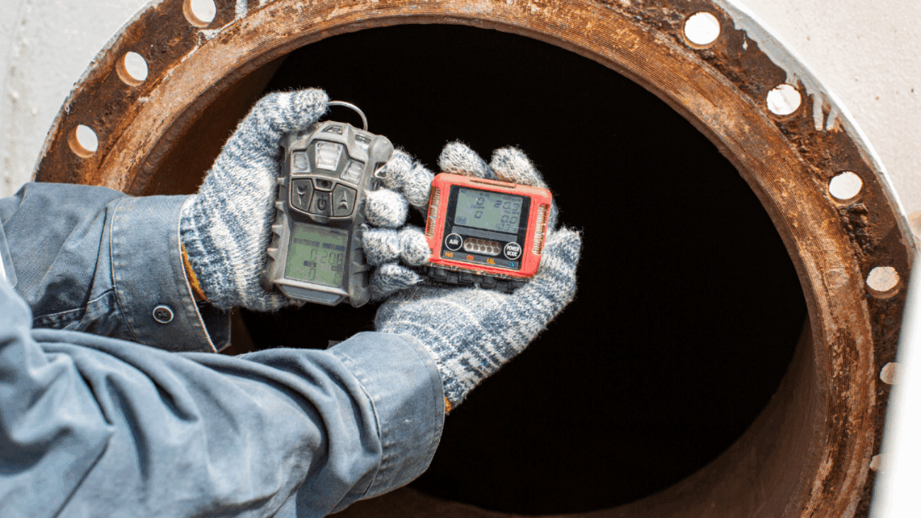 Two hands with gloves on them hold up a gas monitor in front of a confined space, a tunnel