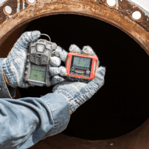 Two hands with gloves on them hold up a gas monitor in front of a confined space, a tunnel
