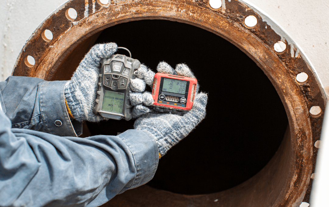 Two hands with gloves on them hold up a gas monitor in front of a confined space, a tunnel